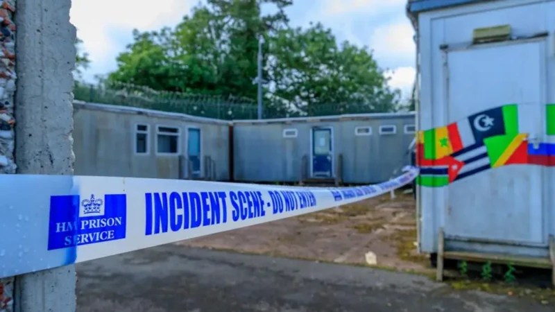Single-storey huts at Campsfield House with flags of various nations painted on the one closest to the camera. There is barbed wire on the roofs of the huts and HM Prison Service blue-and-white tape which says: "Incident scene - do not enter".