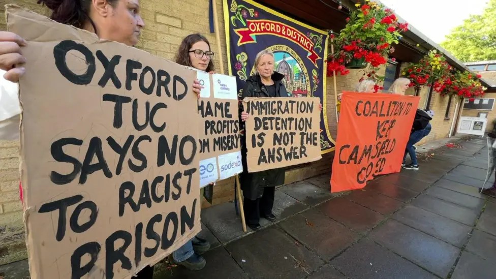 Protesters against the reopening of Campsfield House with placards and the Oxford & District Trades Union banner outside Exeter Hall in Kidlington. Placards have messages including “Oxford TUC says no to racist prison” and “Immigration detention is not the answer”.