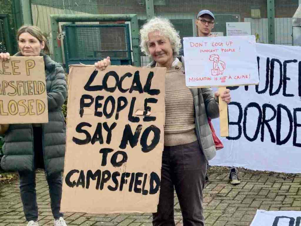 Protesters at the gates of Campsfield House, with campaigner Liz Peretz holding home-made placards reading "Local people say no to Campsfield" and "Don't lock up innocent people. Migration is not a crime." illustrated with a drawing of Paddington Bear.  Image via Coalition to Keep Campsfield Closed