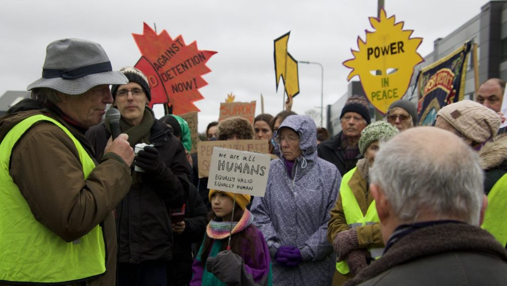 Bill MacKeith addresses a group of protesters outside Campsfield House. Placards carried by protesters include the message "We are all HUMANS. Equally valid. Equally human." (Photo: David Hays for Cherwell)