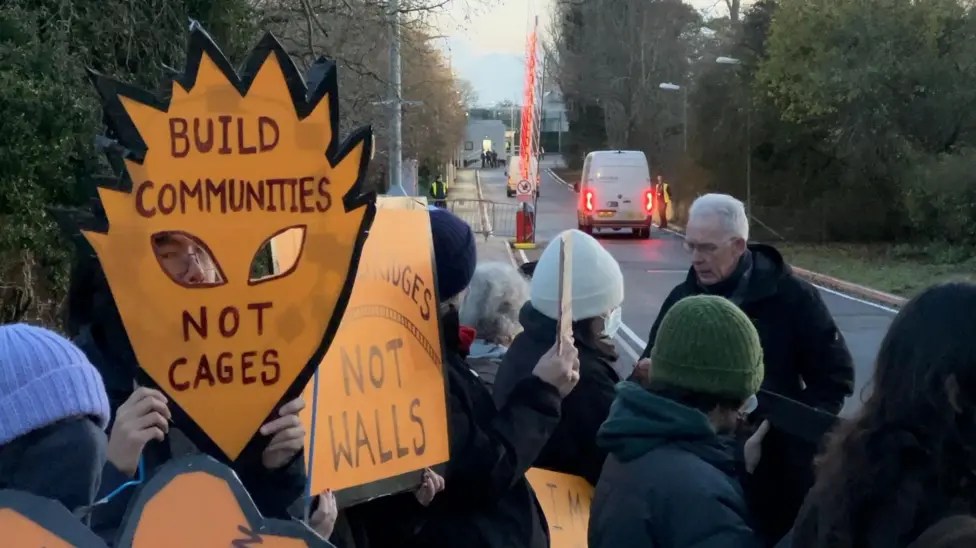 Protesters gather outside the entrance of Campsfield, where a barrier has lifted for a van to pass through. One placard reads: "Build communities, not cages."