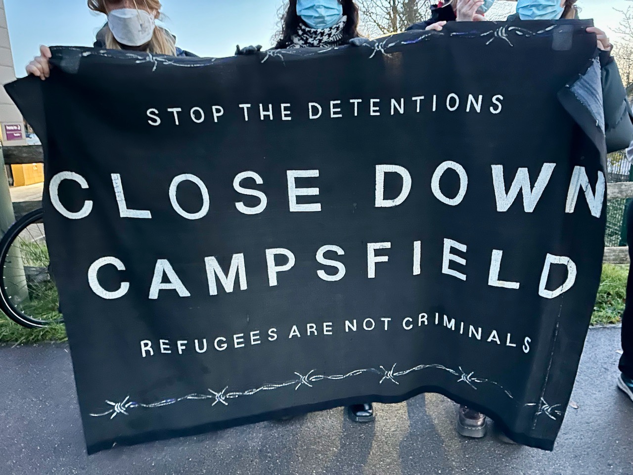 Coalition supporters holding up banner from over a decade ago, reading "Stop the detentions. Close down Campsfield. Refugees are not criminals."