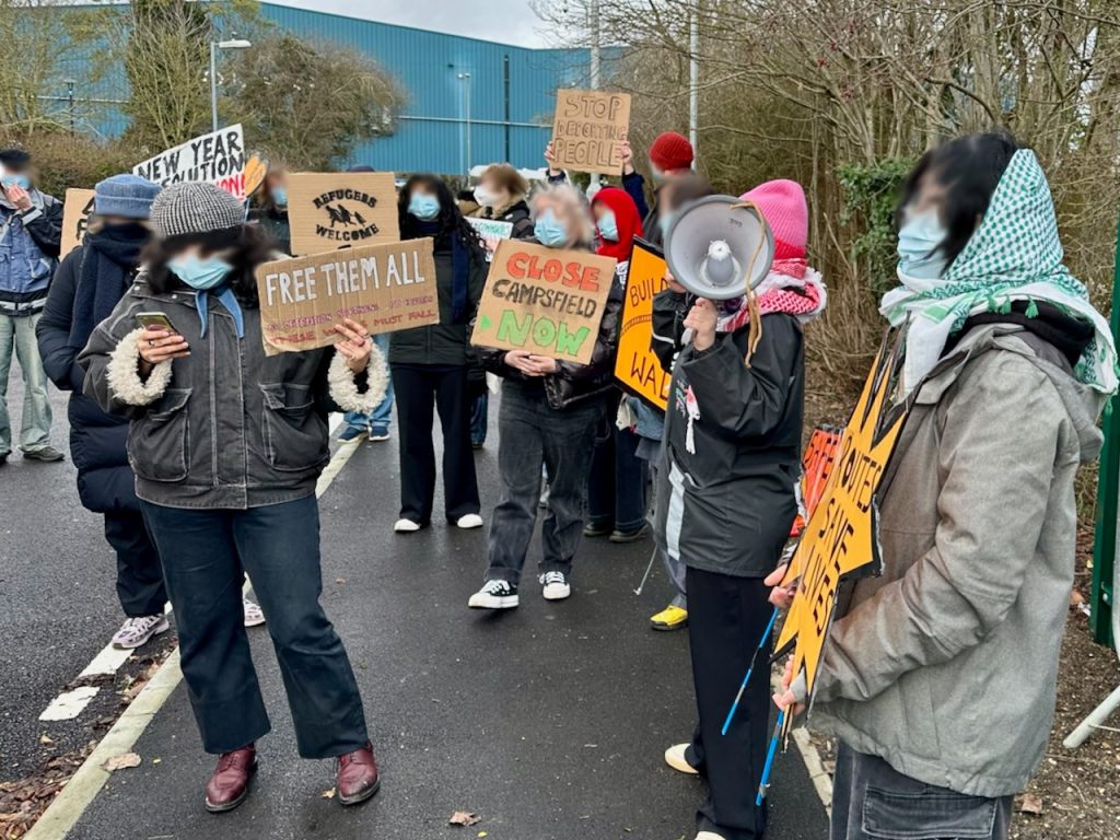 Protestors with signs reading "Refugees welcome", "Free them all", "Stop deporting people" and "Close Campsfield now"