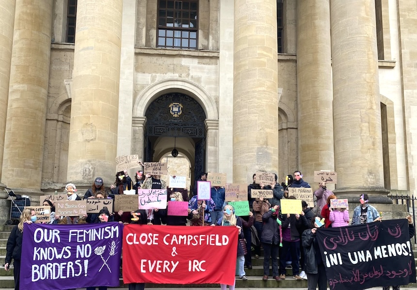 Large crowd gathered for the Feminism Knows No Borders protest at the Clarendon Building, Broad Street, Oxford city centre on 8 March, International Women's Day 2026. Banners at the front include "Our feminism knows no borders!", "Close Campsfield & Every IRC" and a banner, possibly in Persian and Arabic and Spanish, reading "Ni uno menos!" (translated to English: "Not one [woman] less", a "collective scream against machista violence")
